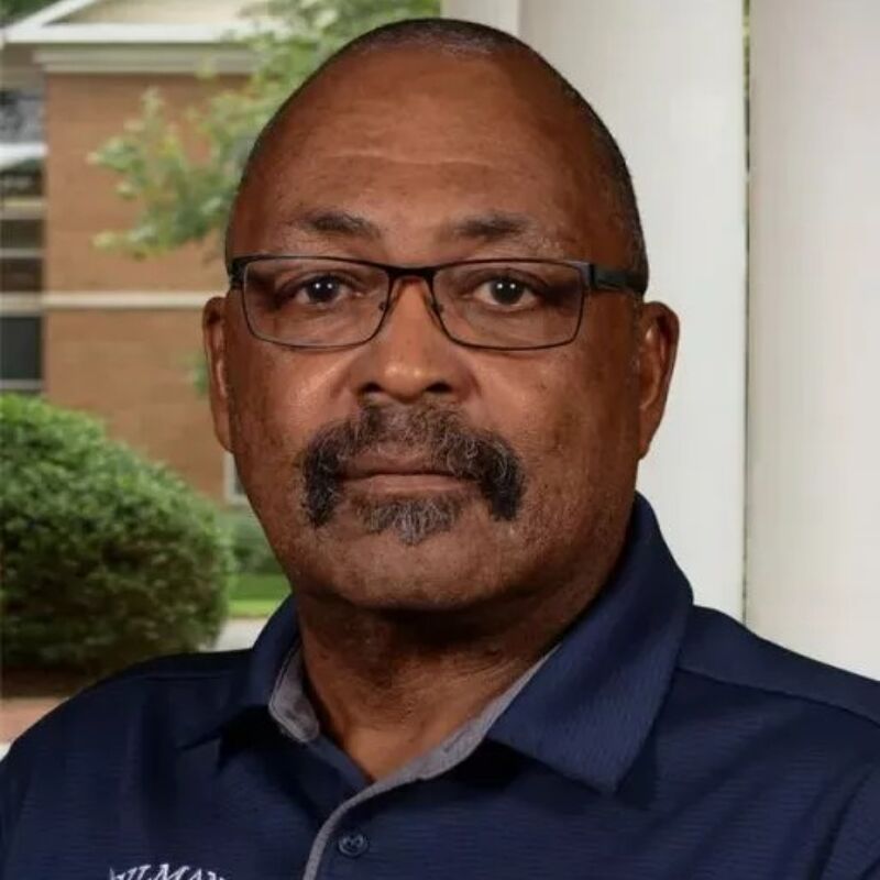 The image shows a close-up of a Black man with glasses and a mustache. He is wearing a dark blue polo shirt. The background is blurred, but it appears to be outdoors, possibly in front of a building. The man has a serious expression.
